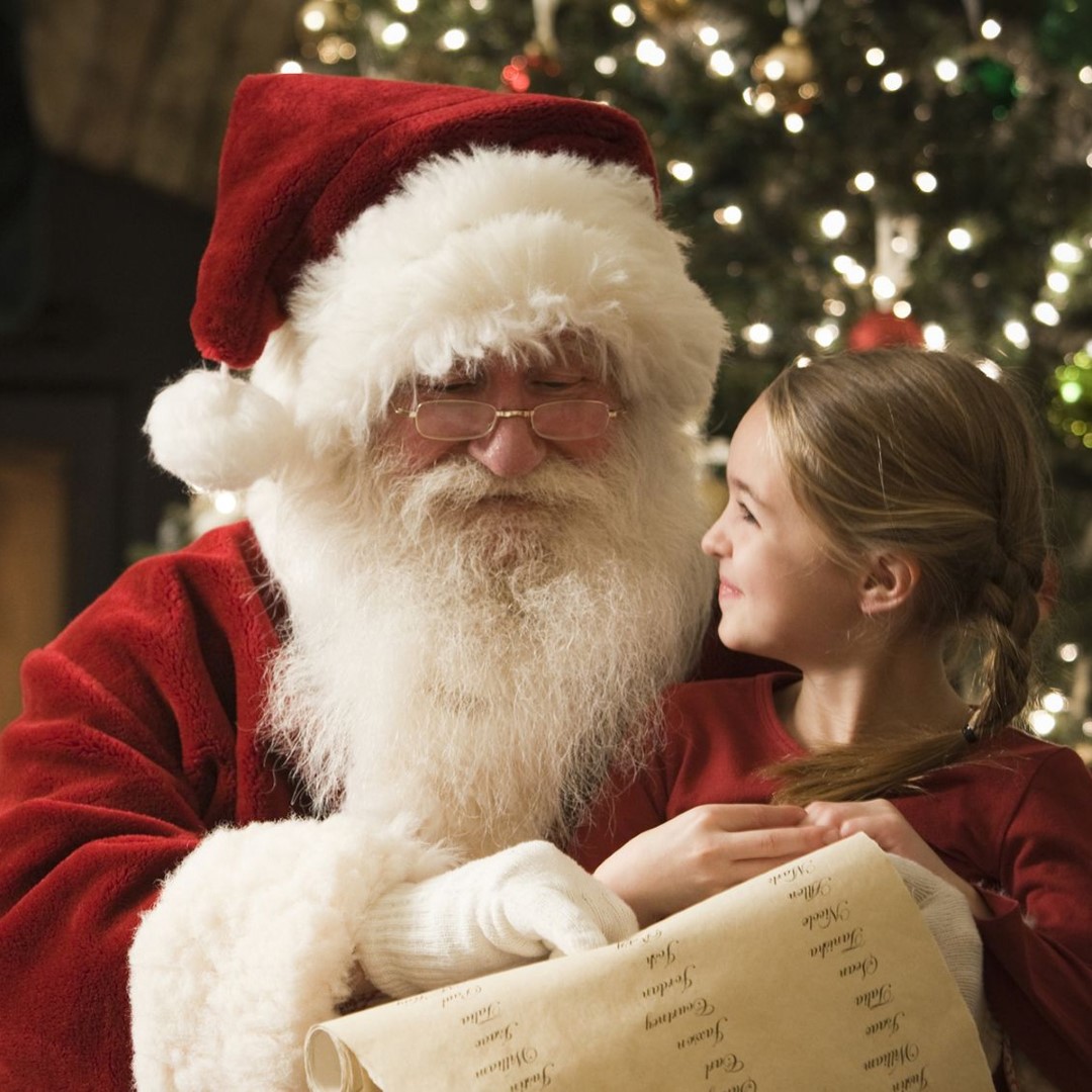 Mikołaj w czerwonym stroju szukający na liście imienia dziewczynki siedzącej  mu na kolanie.